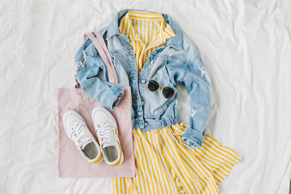 Flat lay image of an outfit including a striped yellow dress, a jean jacket, sneakers, a tote bag, and sunglasses, a great New Orleans Outfit Idea.