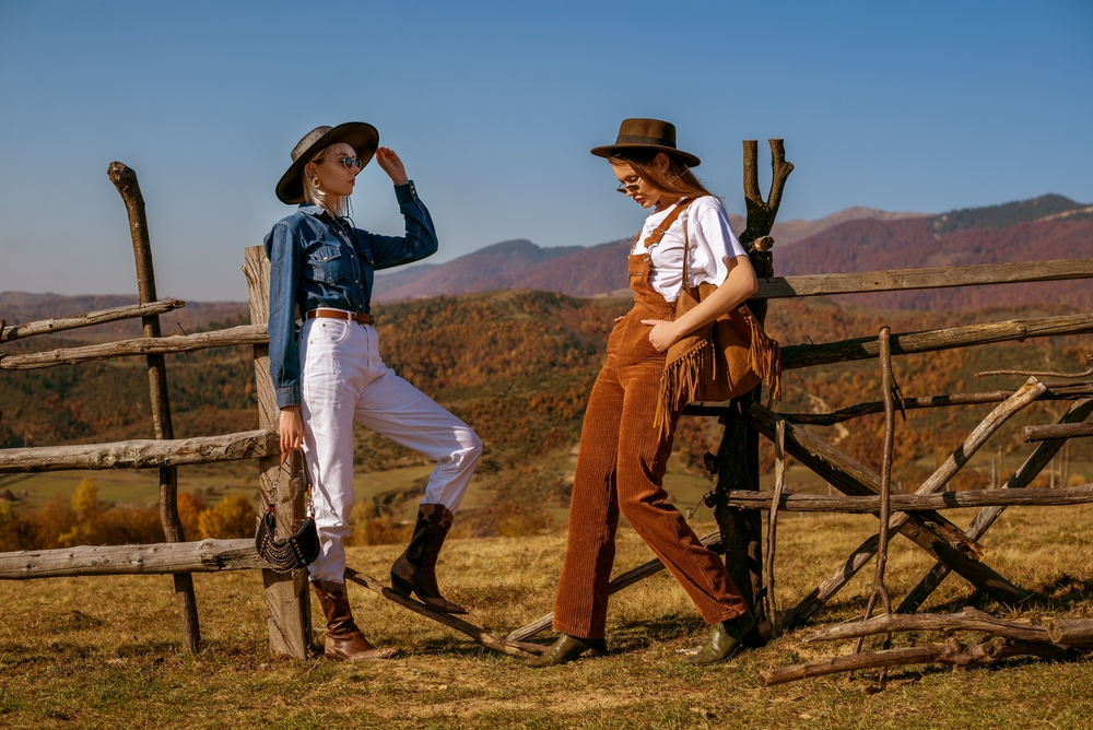 Two women wearing dungarees leaning on a fence. 