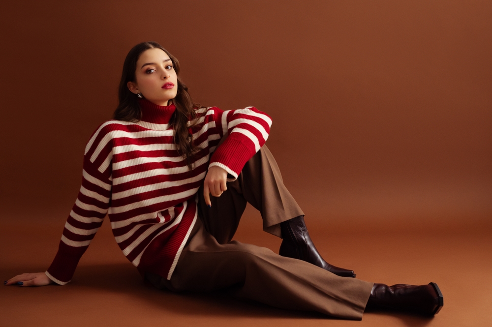 women wearing red striped sweater and brown trousers sat down against plain background. 