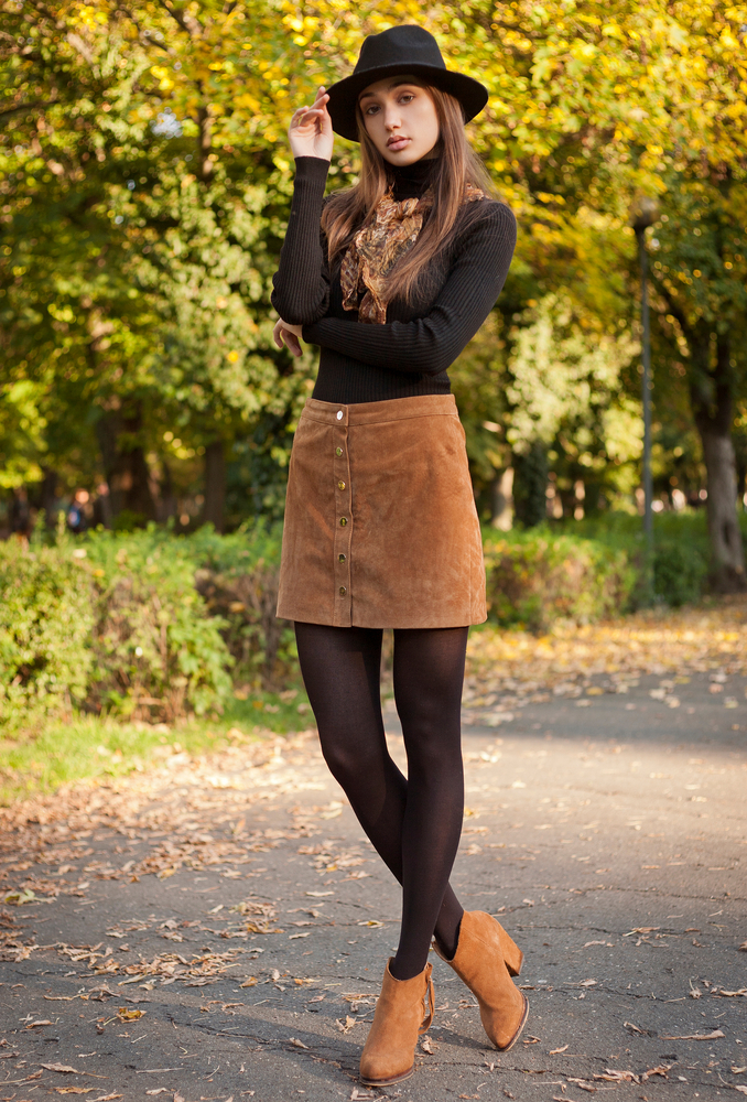 a gil posing in black hat, turtleneck, scarf with brown skirt and booties standing in front of trees