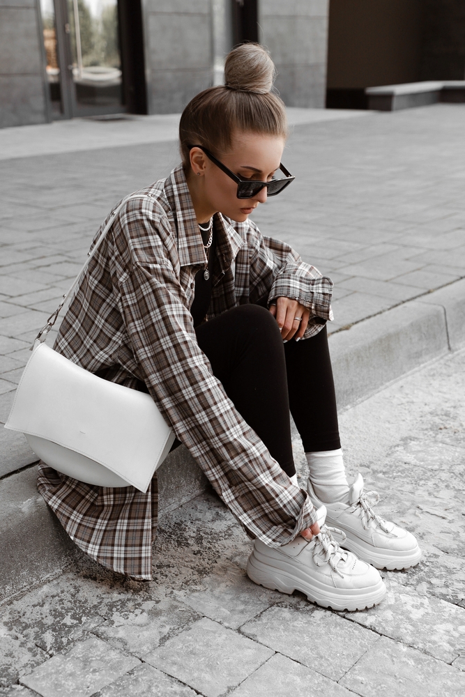 a girl sitting on a street curb with leggings, white sneakers and a long sleeeve flannel shirts