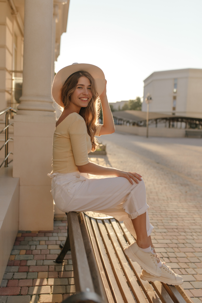 a girl sitting on a park bench with a hat wearing pants and top