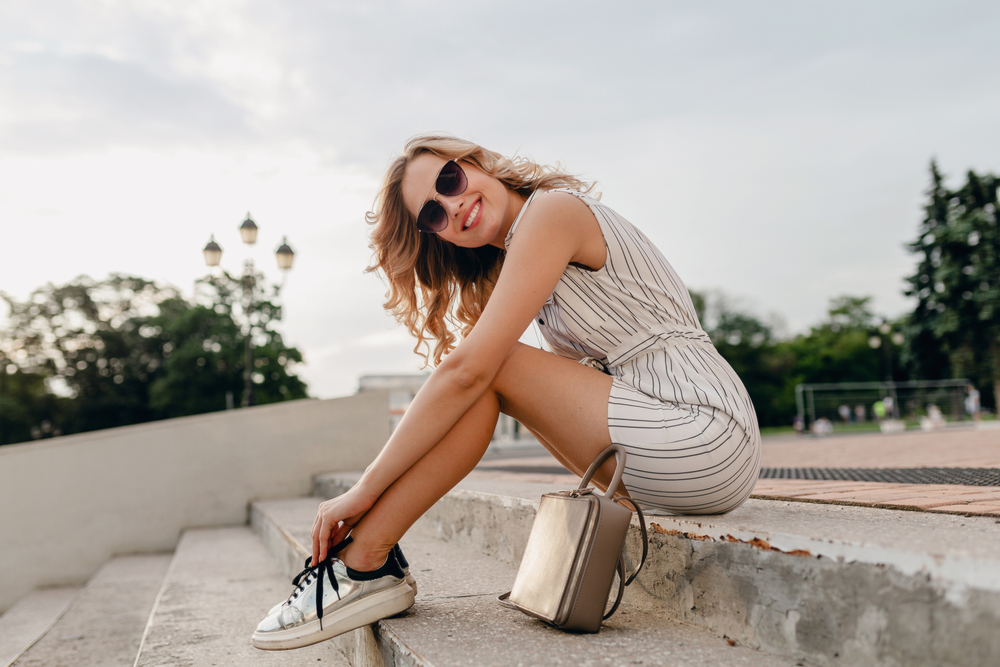 a woman posing on.the steps sitting down in a dress, sneakers and sunglasses