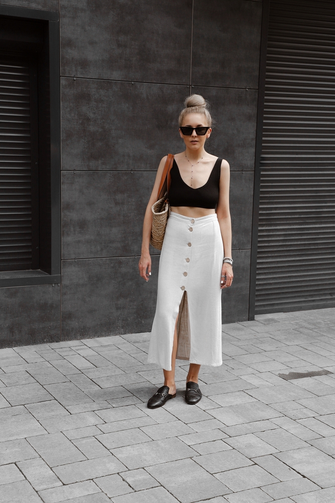 a woman standing on sidewalk in a white linen skirt, black crop top, straw bag and loafers during what to wear in DC during summer