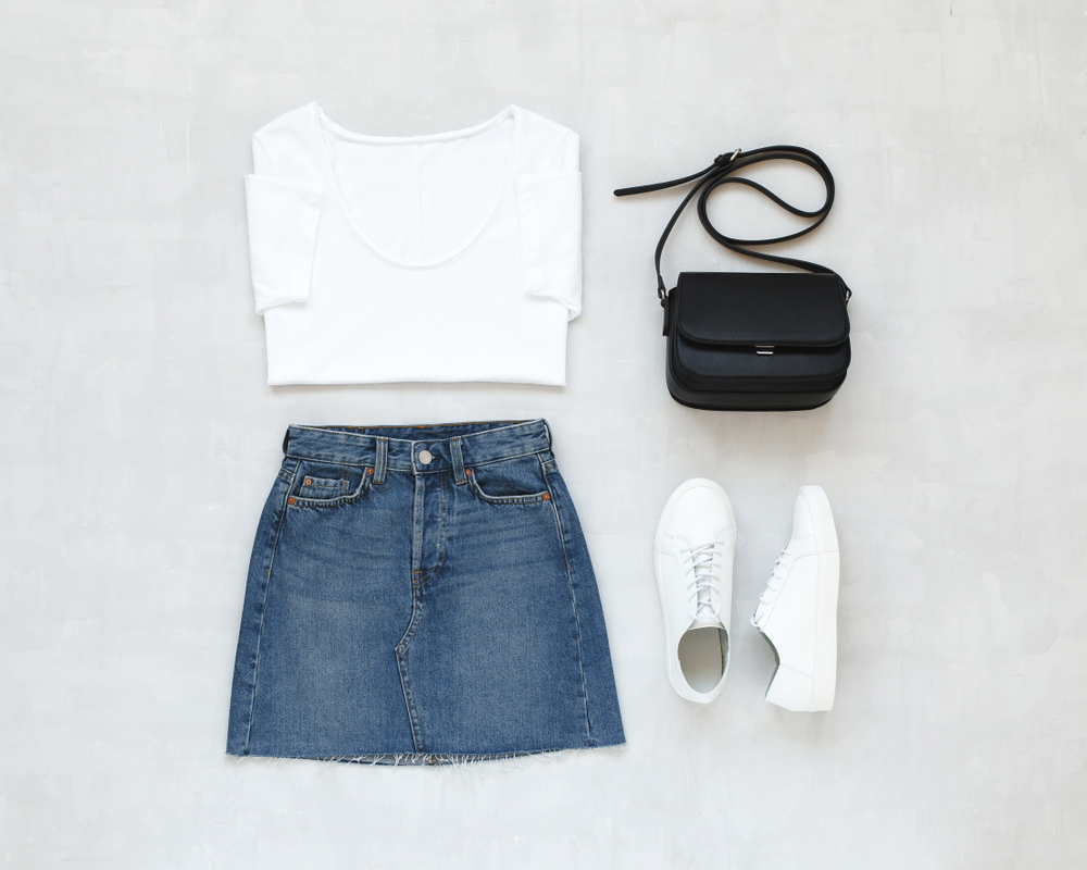 a flat lay of a denim skirt and a white top and white sneakers next to a black cross body bag 