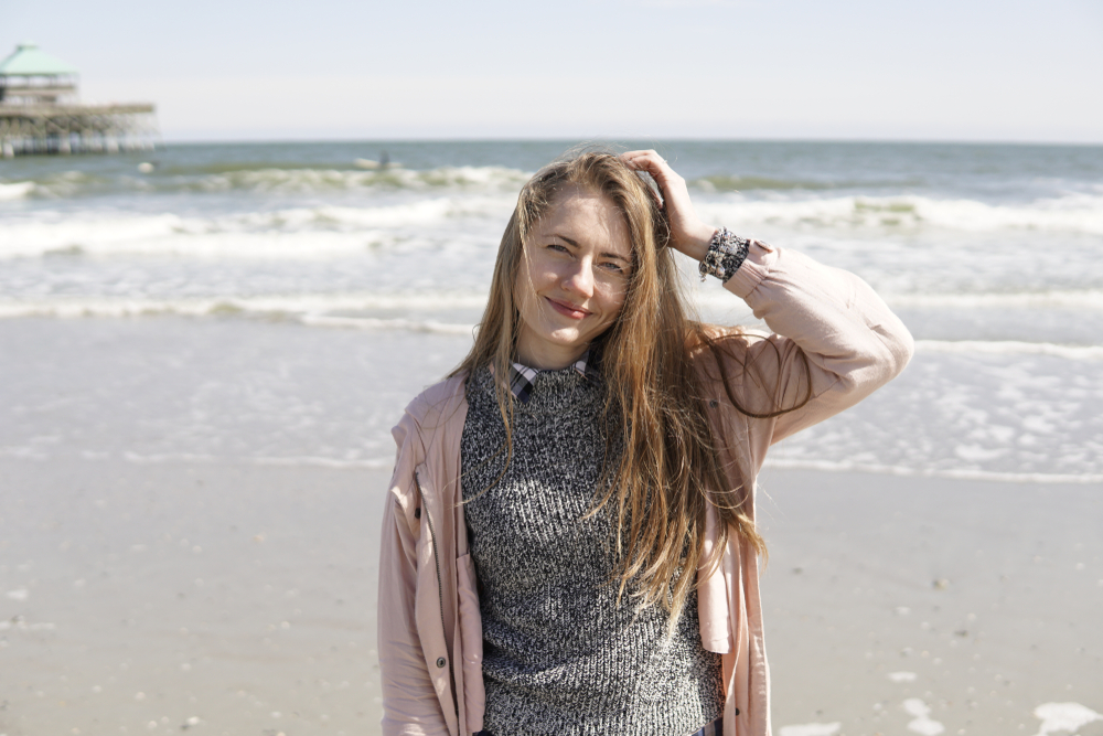 a woman is standing on the beach with a collared shirt under a sweater under a rain jacket, a winter charleston sc outfit idea 