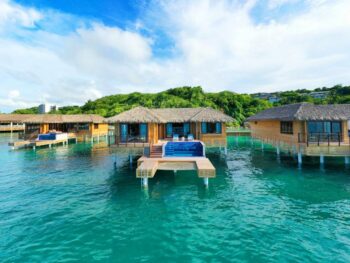 overwater bungalow with thatched roof and pool standing in blue water with blue sky and clouds. the building is brown