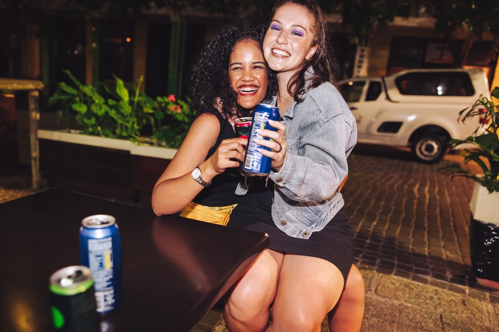 Two friends sit on one another's laps, enjoying a night out in Atlanta in their black dresses, drinks in their hands, knowing what to wear in Atlanta on evenings out.