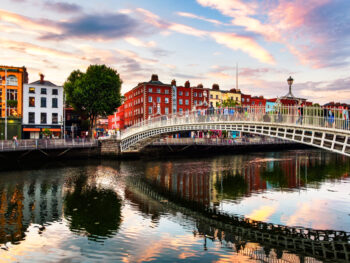As the sun sets, a photo of a bridge in Dublin connects to sides of the streets over a river, tall buildings and pubs around