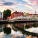 As the sun sets, a photo of a bridge in Dublin connects to sides of the streets over a river, tall buildings and pubs around
