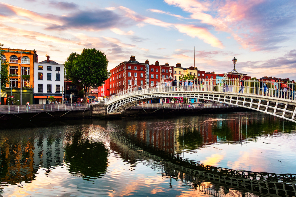 As the sun sets, a photo of a bridge in Dublin connects to sides of the streets over a river, tall buildings and pubs around: this is one of the cheapest places to fly from Atlanta. 