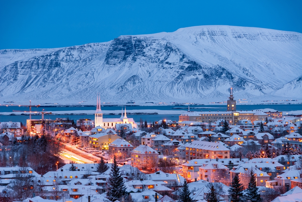 An overhead shot of Reykjavik shows the warm glow of city lights at night in the winter, mountains snow capped beyond it. 