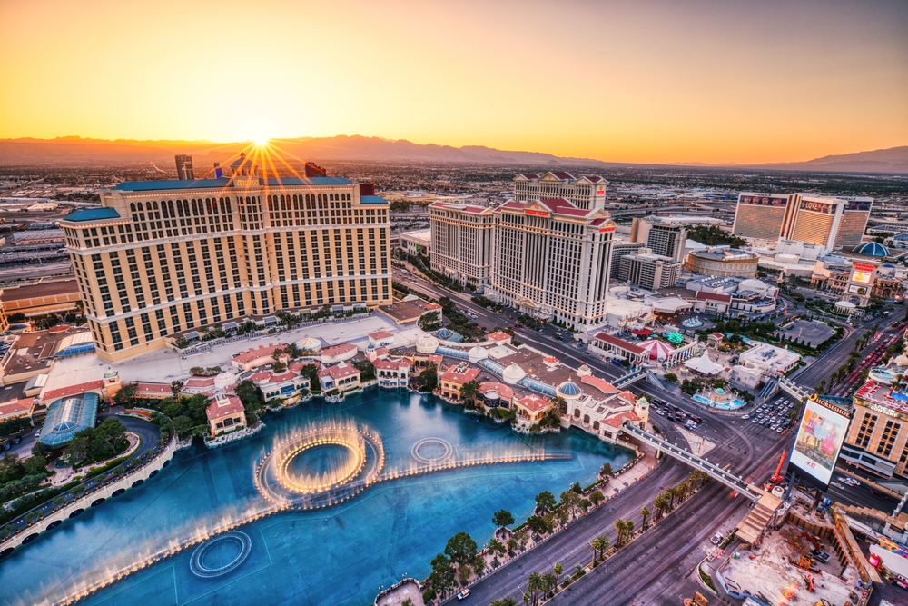 An overhead shot of Vegas shows a strip alive with hotels, fountains, streets, mountains in the back and the sun rising. 