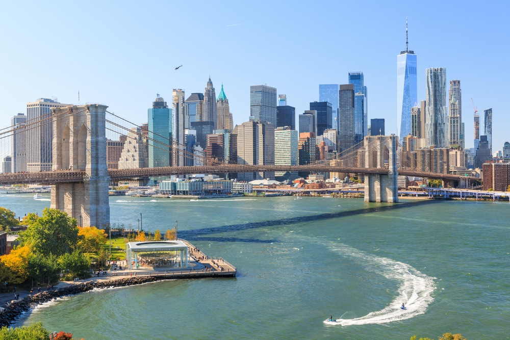 A skyline shot of New York City, which just so happens to be one of the cheapest places to fly from Dallas, features the Brooklyn Bridge over the river, skyscrapers in the background.