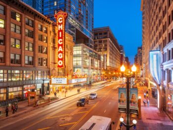A street in Chicago is lit up at night with street signs a glow as cars travel down the road. the sky is blue but it is dusk and there is a sign that is red and say the word "chicago"