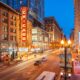 A street in Chicago is lit up at night with street signs a glow as cars travel down the road. the sky is blue but it is dusk and there is a sign that is red and say the word "chicago"
