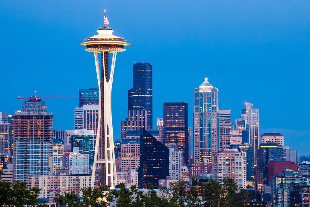 Seattle features the iconic Space Needle amongst a series of tall buildings and skyscrapers, their lights turning on at dusk. 
