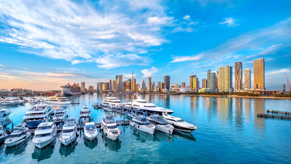 A shot of Miami, one of the cheapest places to fly from Washington D.C., shows a boat harbor, the blue waters, and the tall building's on the coast.