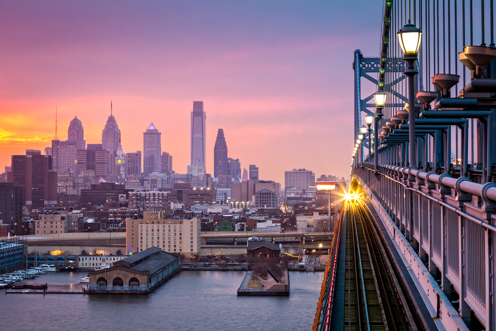 The sun begins to set in Philly, one of the cheapest places to fly from Washington D.C., its light reflecting off a bridge that connects over the water, the skyline in the back.