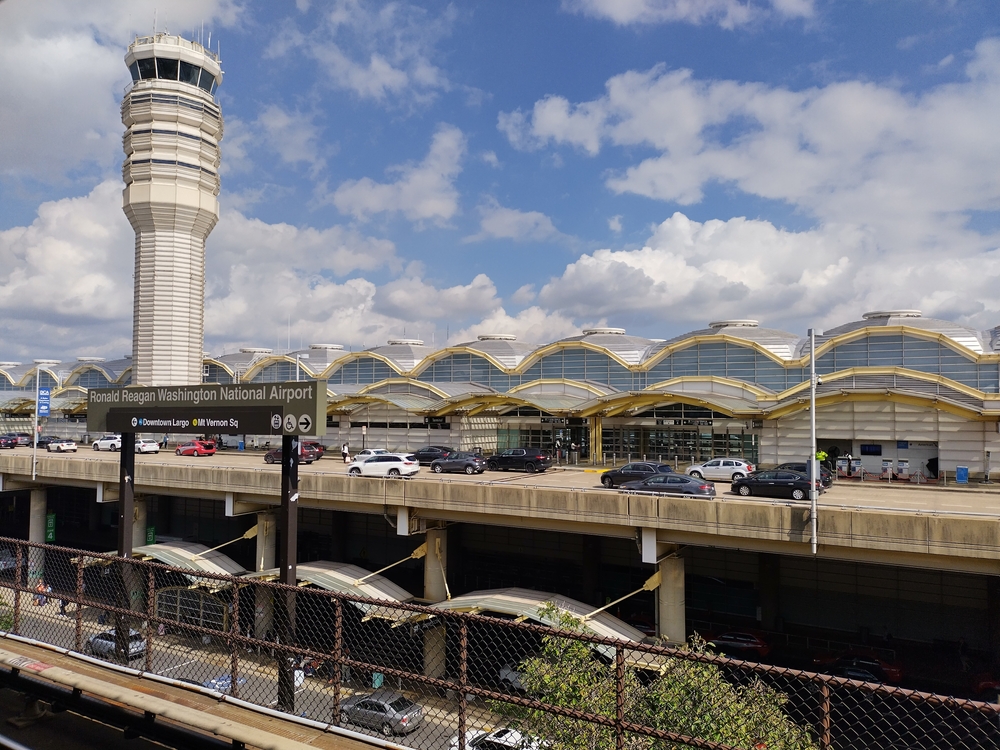 A shot of the Washington DC airport shows cars puling up to a building with curved roofs and flight towers, with the national airport sign on the front! People visiting know that there are some of the cheapest places to fly from Washington D.C., so they fly out from here.