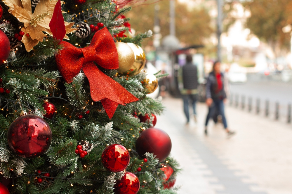 Green spruce tree with red decorations, bell, bow. People out of focus. 
