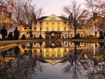 The Williamsburg Inn decorated for the holidays is shown reflected in an outdoor fountain. the building is lit in gold and yellow christmas lights and flanked by two dead trees on either side with a pond in the foreground