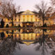 The Williamsburg Inn decorated for the holidays is shown reflected in an outdoor fountain. the building is lit in gold and yellow christmas lights and flanked by two dead trees on either side with a pond in the foreground
