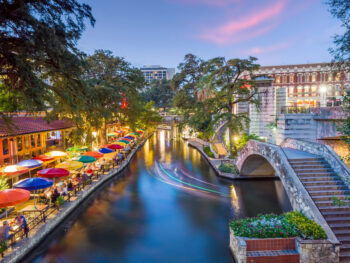 River walk in San Antonio city downtown skyline cityscape of Texas USA at sunset. there are colorful umbrellas on the left of the photo and water in the middle and a bridge to the right.