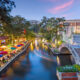 River walk in San Antonio city downtown skyline cityscape of Texas USA at sunset. there are colorful umbrellas on the left of the photo and water in the middle and a bridge to the right.